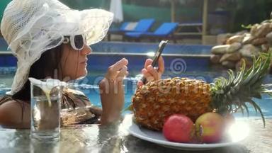 年轻漂亮的女人在游泳池里用手机，在盘子旁边喝水，吃热带水果。 3840x2160